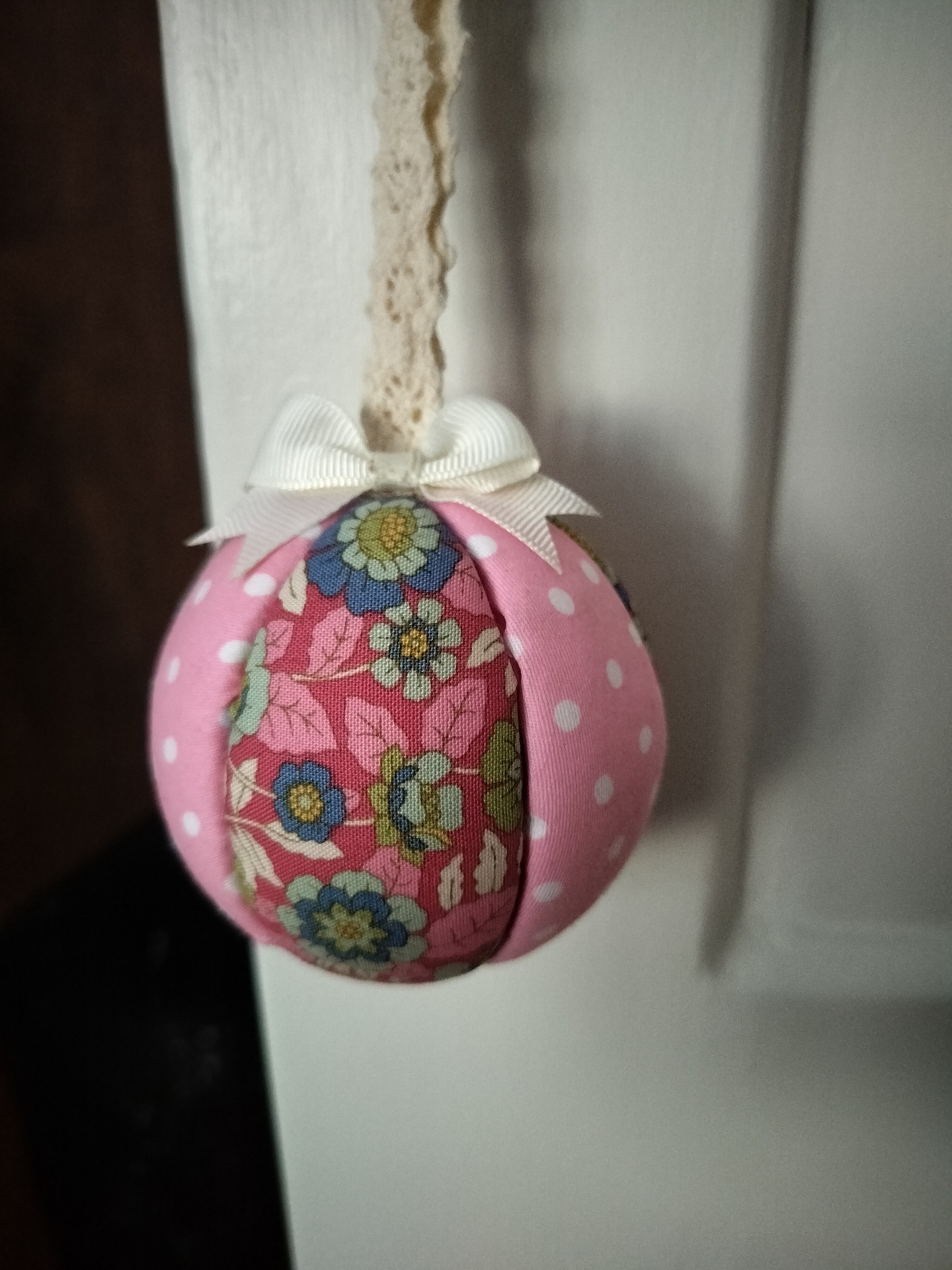 Vaaleanpunainen styroxpallo, jossa kukkakuvio ja valkoinen pitsinauha, roikkuu valkoisessa taustassa.
